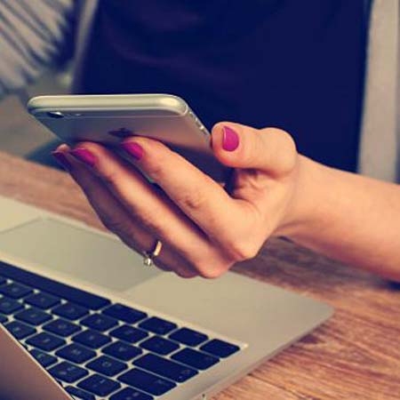 Closeup of a Woman's Hand Who is Ordering Cannabis Delivery On Her Mobile Phone Sitting in Front of a Laptop on a Desk